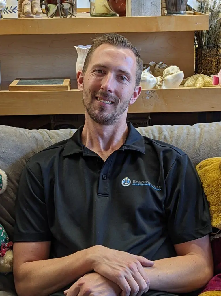 A man with short light brown hair and a trimmed beard sits on a gray couch, smiling. He is wearing a black polo shirt with a blue logo. Shelves with decorative items and plush toys are in the background.