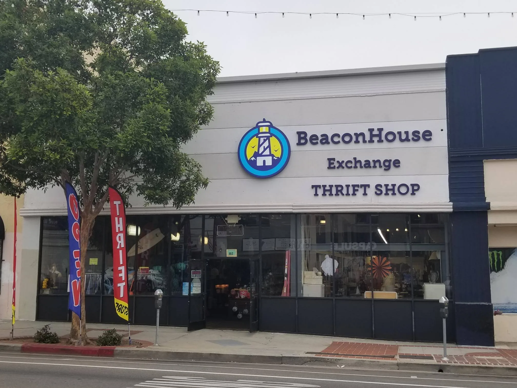 A storefront with a sign reading BeaconHouse Exchange Thrift Shop above large windows. A tree stands in front, and a red vertical flag with THRIFT is on the left side by the sidewalk.