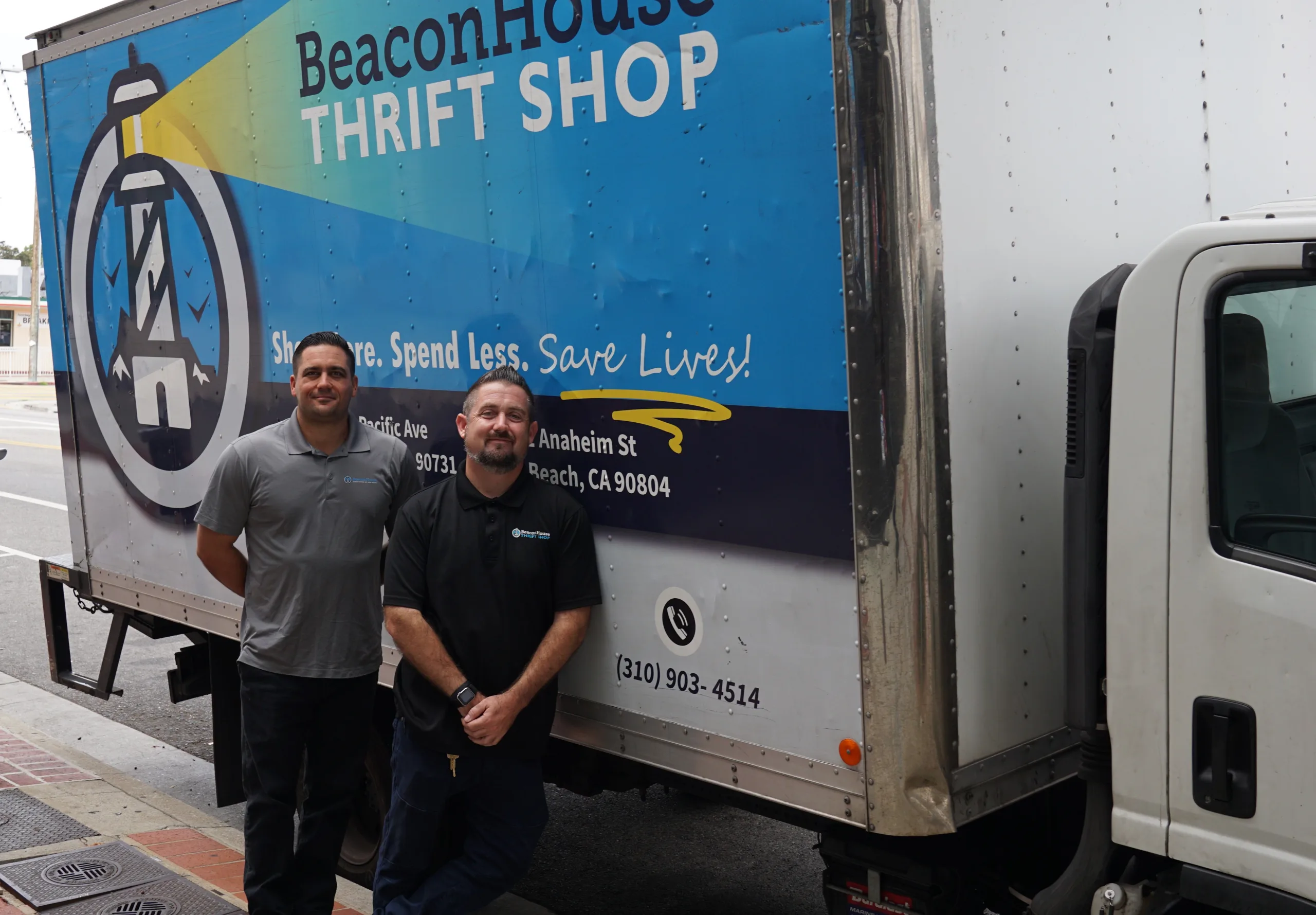 Two men stand and smile in front of a Beacon House Thrift Shop truck parked on a street. The truck features the shop’s name, logo, address, phone number, and colorful graphics.