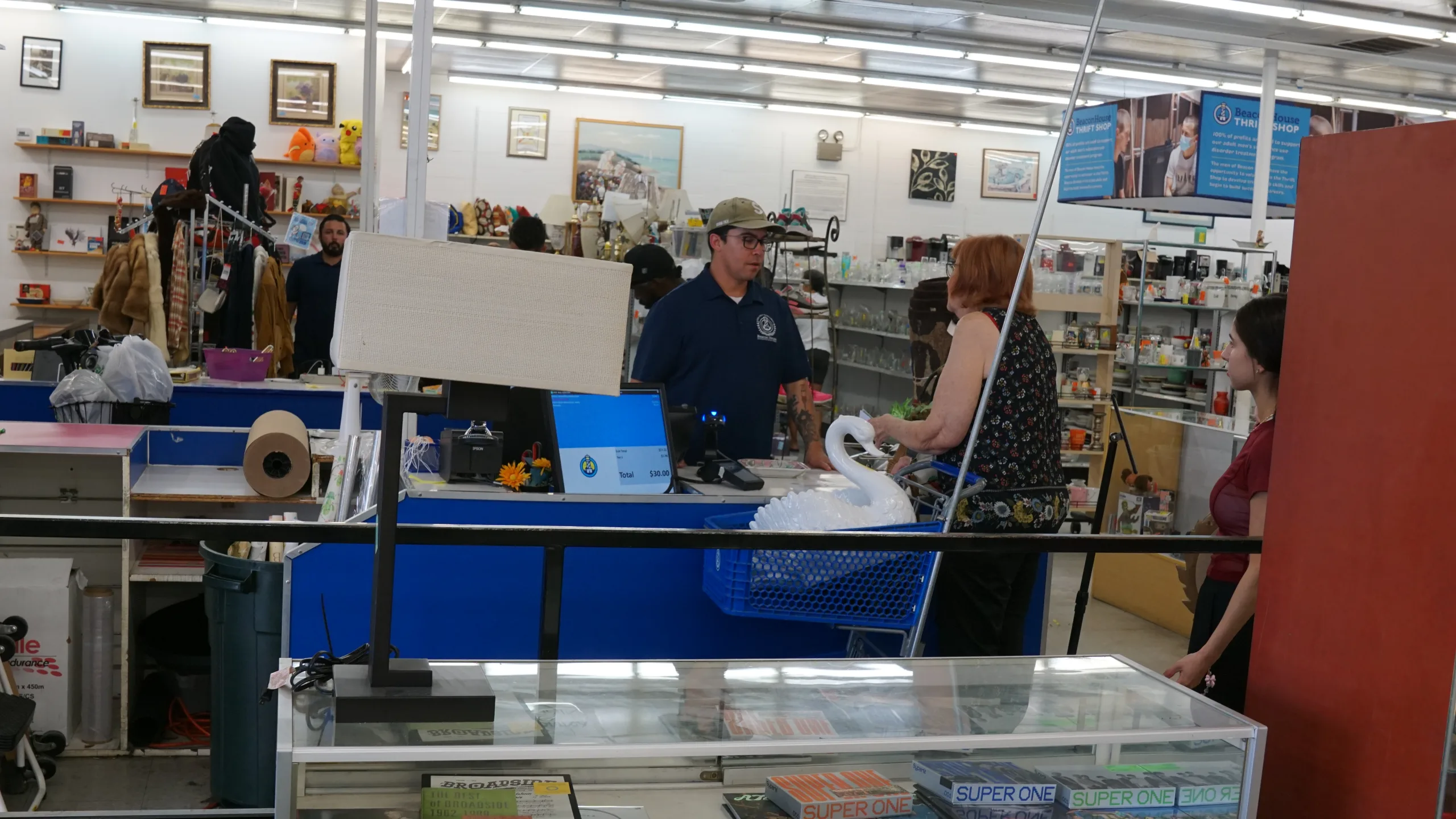 A thrift store checkout area with a man in a blue shirt at the register assisting two women, one holding a shopping basket with a white swan item. Shelves of assorted goods and a glass display case are visible.