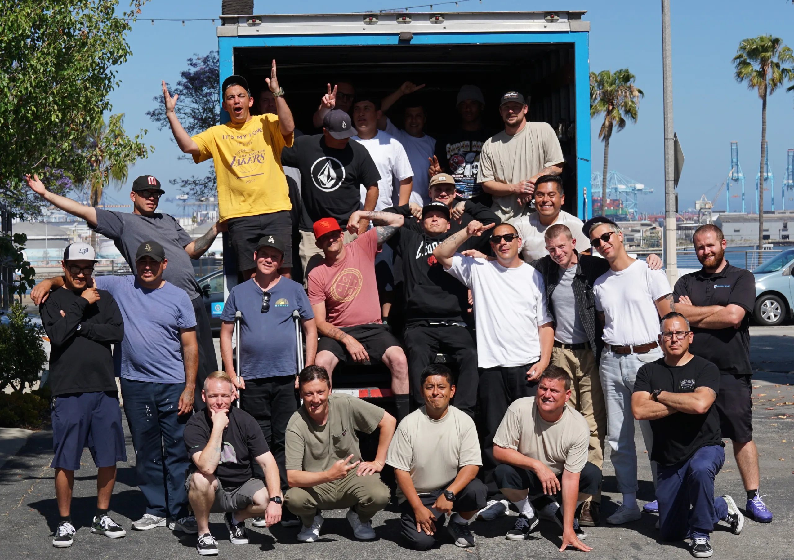 A group of men posing playfully and smiling in and around a large open truck on a sunny day, with palm trees and industrial cranes visible in the background.