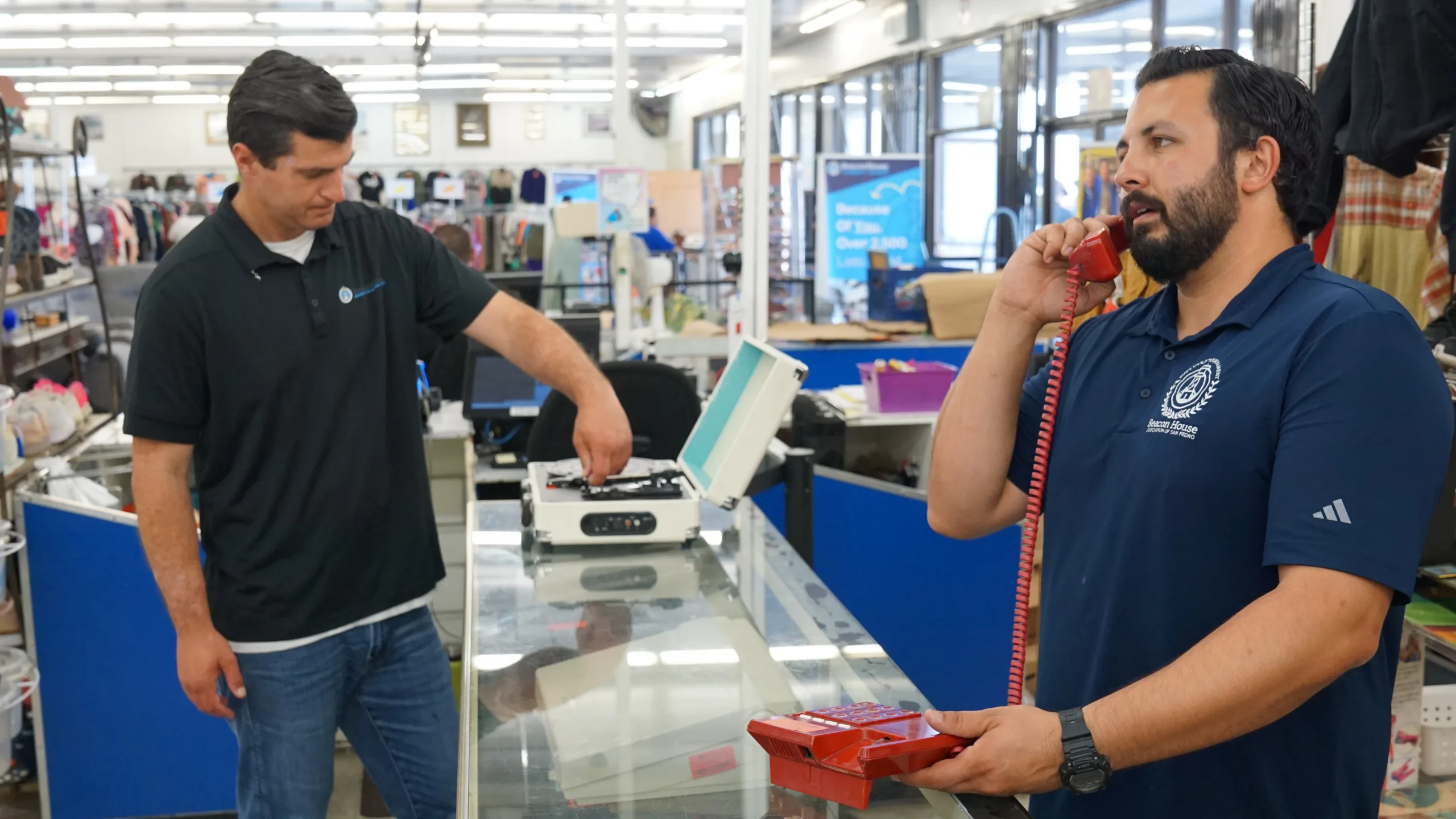 Two men stand behind a glass display counter in a store. One uses a red telephone while the other adjusts a turntable. The store has bright lights, blue counters, and shelves of assorted items in the background.