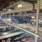 A thrift store interior with shelves of toys, racks of clothes, and a large sign above reading “Shop More. Spend Less. Save Lives!” An American flag hangs nearby, and exposed ceiling beams are visible overhead.