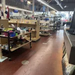 A thrift store interior with shelves displaying assorted household items, dishes, and baskets. The store has high ceilings, exposed beams, large windows, and flags hanging above the shelves.