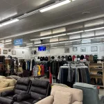 A thrift store interior with couches and armchairs in the foreground and racks of men’s clothing in the background. A blue sign labeled “Men’s Clothing” hangs from the ceiling above the racks.