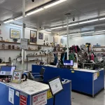 A thrift store interior with blue counters, computers, and No refunds or exchanges signs. Shelves on the walls display clothes, toys, and various items. The store is well-lit with fluorescent lights.