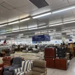 A thrift store interior with various couches, armchairs, dressers, and other furniture items displayed. Overhead fluorescent lights illuminate the space; a blue sign in the background reads, Shop More. Spend Less. Save Lives.