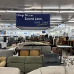A thrift store interior with sofas, chairs, and furniture. A large hanging sign reads, Shop More. Spend Less. Save Lives! The store is well-lit and filled with various home goods.