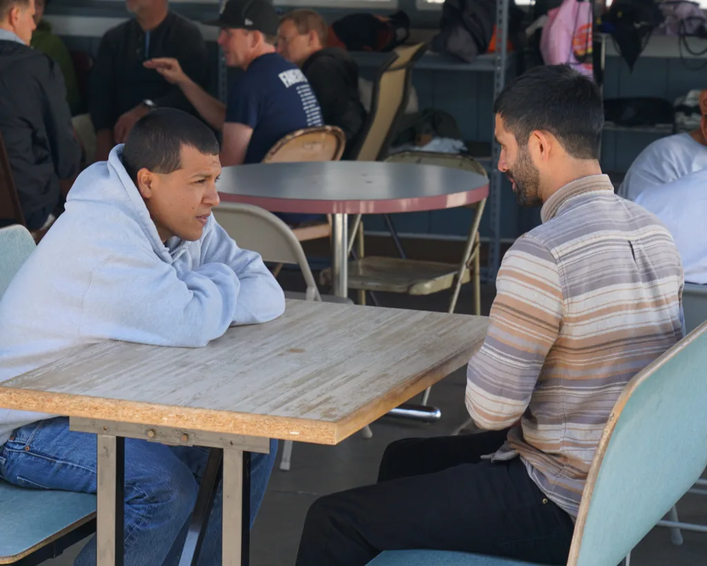 Two men sit across from each other at a wooden table, engaged in conversation. Other people are seated at tables in the background in a casual, indoor setting.