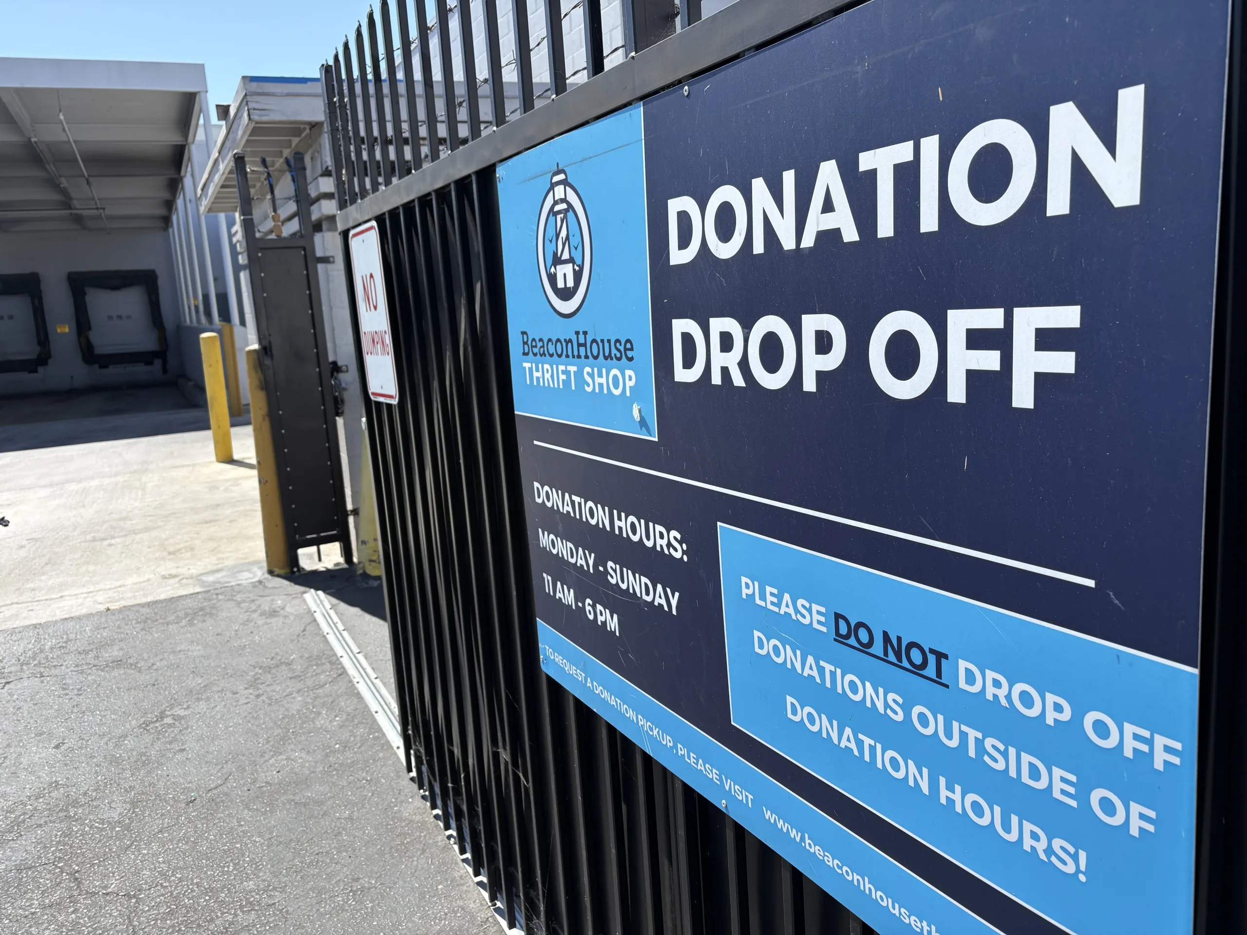 A large sign on a black gate reads Beacon House Thrift Shop Donation Drop Off. Donation hours are listed as Monday-Sunday, 11am-6pm. A warehouse and loading docks are visible in the background.
