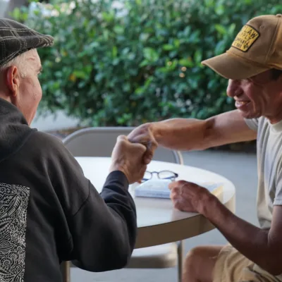 Two men sit at a round outdoor table, smiling and holding hands in a friendly gesture. One wears a black cap and patterned hoodie, the other a tan cap and t-shirt. Green bushes are visible in the background.