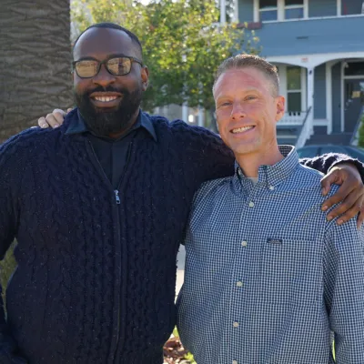 Two men stand outdoors smiling with arms around each other’s shoulders. One wears glasses and a navy sweater, the other wears a blue checkered shirt. Sunlight and trees are visible in the background.